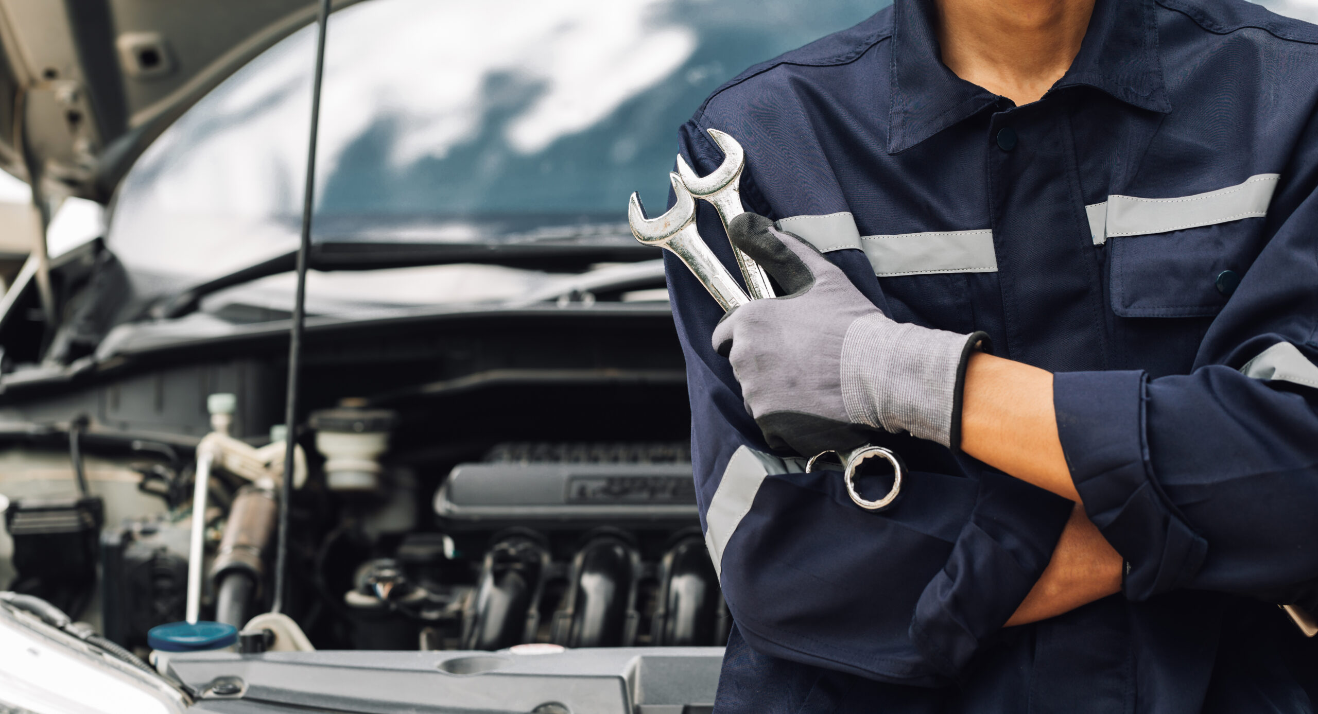 Mechanic holding wrenches in front of a car with the hood open, representing rent vs. buy options for vehicle repair tools.